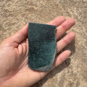 May include: A rectangular slab of dark forest green stone with a smooth, polished surface and a rough, unpolished gray edge at the bottom. The stone features subtle mottled patterns and variations in color throughout its deep green surface.