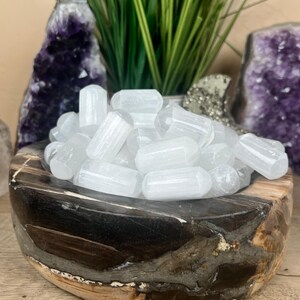 May include: A wooden bowl filled with white selenite crystals. The bowl has a dark, multi-toned finish. Green grass and purple amethyst crystals are in the background. The crystals are cylindrical and translucent.