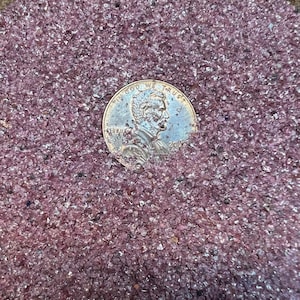 Puede incluir: Primer plano de una moneda de un centavo de dólar estadounidense con la inscripción "In God We Trust" y "Liberty" en el anverso. La moneda está apoyada sobre un lecho de arena rosa y brillante.