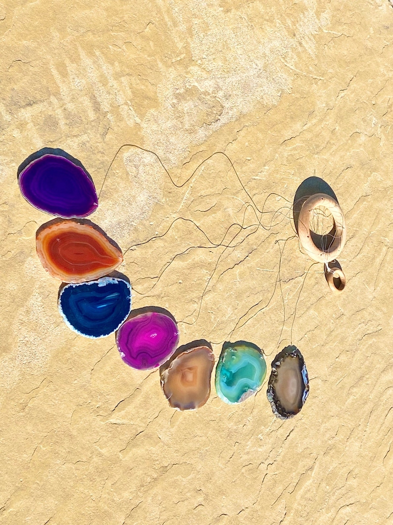 May include: A wind chime made of agate slices in various colors, including purple, red, blue, pink, brown, and green. The slices are suspended from a wooden ring.