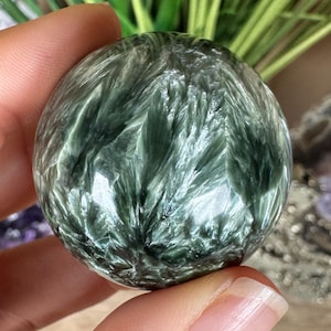 Puede incluir: Una piedra preciosa de serafinita pulida en forma de esfera, con una base de color verde bosque profundo e inclusiones plateadas y blancas complejas, plumosas y chatoyantes que crean un patrón radiante sobre la superficie lisa.