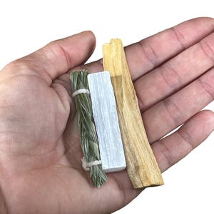May include: A set of three smudging sticks, including white sage, palo santo, and a white selenite stick. The sticks are bundled together in a hand.