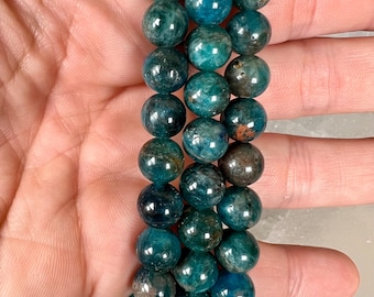 May include: A stack of teal-colored gemstone bead bracelets. The beads are round and have a polished, glossy finish. The color varies from deep teal to lighter turquoise, with some beads showing brown and gray inclusions. The bracelets are displayed on a white surface.