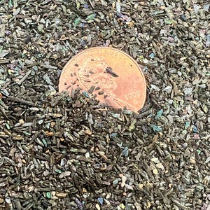May include: A close-up of a pile of iridescent mica flakes, with a penny for scale. The flakes are a mix of dark green, black, and silver, with a slight shimmer.