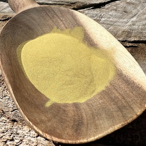 May include: A wooden scoop filled with a pile of fine, yellow powder. The scoop is made of dark wood with a handle and a wide, shallow bowl. The powder is a vibrant yellow color and appears to be finely ground. The scoop is resting on a weathered wooden surface.