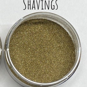 May include: A small clear container filled with gold glitter. The container has a silver lid and the word "SHAVINGS" is written above the container.