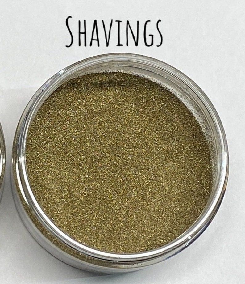 May include: A small clear container filled with gold glitter. The container has a silver lid and the word "SHAVINGS" is written above the container.