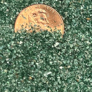 May include: A close-up of a penny, partially submerged in a bed of green glass beads. The penny features the inscription "In God We Trust".