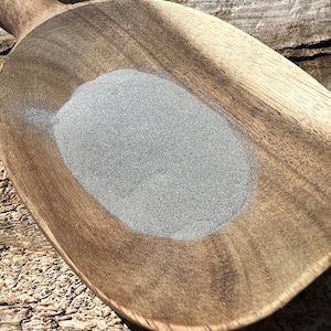 May include: A wooden scoop filled with a pile of fine, gray powder. The scoop is made of light brown wood with a handle. The powder is a uniform color and texture, contrasting with the wood's grain.