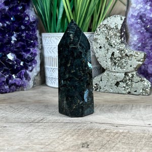 Pode incluir: Uma ponta de cristal verde escuro e preto com uma ponta pontiaguda. O cristal está sentado em uma superfície de madeira.