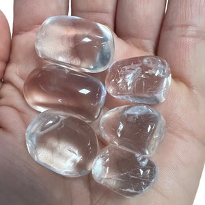 May include: Six clear quartz tumbled stones, also known as rock crystals, in various sizes and shapes. The stones are smooth and polished, with a translucent appearance.