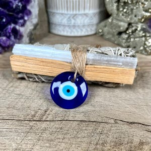 May include: A blue Evil Eye pendant with a white and black eye design, hanging from a bundle of sage and selenite. The pendant is round and has a glossy finish. The sage and selenite are tied together with twine.