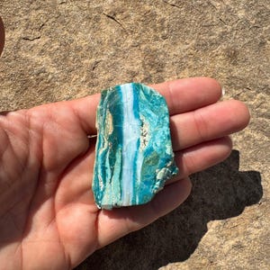 May include: A raw, unpolished mineral specimen featuring vibrant shades of turquoise and teal with a distinct, vertical white band running through the center. The stone has a rough, textured surface with natural variations in color and mineral deposits.