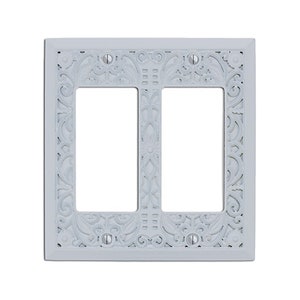 May include: White double-gang wall plate with an ornate, decorative design. The plate has a cutout for two standard electrical outlets.