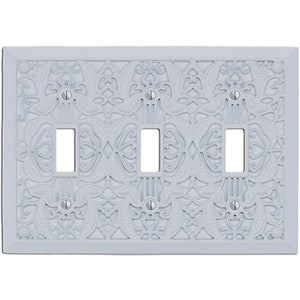 May include: White decorative wall plate with an intricate, embossed design. The plate has three openings for light switches.