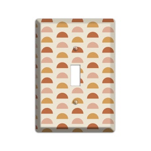 May include: A light switch cover with a geometric pattern of overlapping half circles in shades of pink, orange, and brown on a cream background.