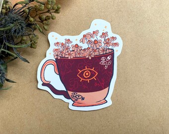 May include: A dark red teacup with a white rim and a single large eye design. The cup is filled with pink and white flowers and has a white handle.