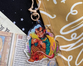 May include: A clear acrylic charm featuring a cartoon character with red skin, horns, and a tail, holding a cup. The charm has a gold-colored clasp. The character is surrounded by stars, a crescent moon, and lavender. The background includes a black fabric with white stars.