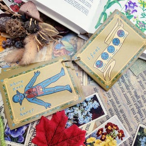 May include: Two illustrated cards with a yellow background and a gold border. The first card shows a blue cartoon figure with a skeleton and internal organs. The text on the card reads "where does it hurt?" The second card shows a banner with a series of blue cartoon faces with different expressions. The text on the card reads "i feel..."