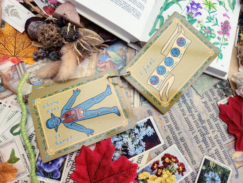 May include: Two illustrated cards with a yellow background and a gold border. The first card shows a blue cartoon figure with a skeleton and internal organs. The text on the card reads "where does it hurt?" The second card shows a banner with a series of blue cartoon faces with different expressions. The text on the card reads "i feel..."