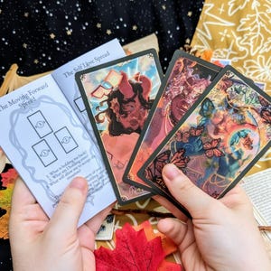 May include: A tarot card reading spread with three cards laid out on a white booklet with the text "The Moving Forward Spread" and the instructions "1. What is holding me back? 2. What am I holding onto? What will allow me to accept this moment?"  The booklet is on a dark background with a fall-themed backdrop.  The cards are from a deck with colorful illustrations.