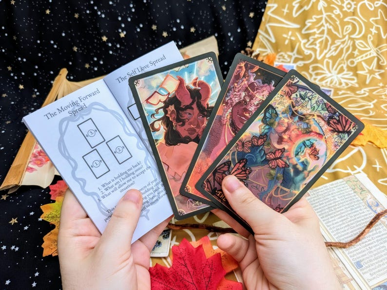 May include: A tarot card reading spread with three cards laid out on a white booklet with the text "The Moving Forward Spread" and the instructions "1. What is holding me back? 2. What am I holding onto? What will allow me to accept this moment?"  The booklet is on a dark background with a fall-themed backdrop.  The cards are from a deck with colorful illustrations.
