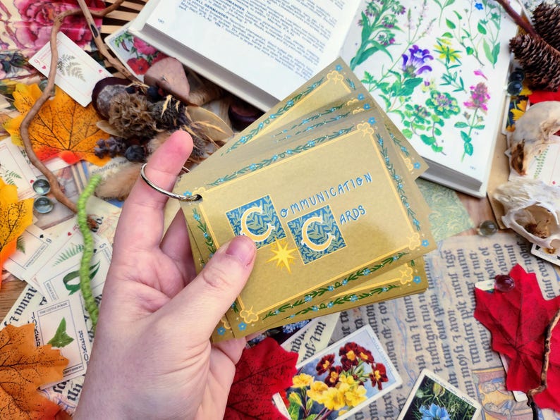 May include: A set of gold communication cards with blue accents and the text "Communication Cards" printed on the front. The cards are held in a hand.