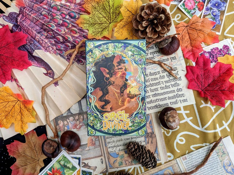 May include: A tarot card with a green, blue, and gold border. The card features a woman with pointed ears and long brown hair. She is holding a glowing orb in her hand. The card is titled "Oracle Diviner".