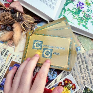 May include: A set of gold-colored communication cards with a blue and gold logo that says "Communication Cards". The cards are fanned out and held by a hand.