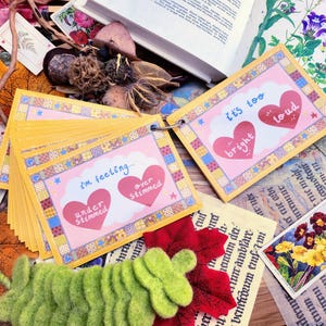 May include: A set of colorful cards with different phrases about feelings, including "I'm feeling... under stimulated, over stimulated", and "It's too bright, it's too loud". The cards are arranged in a stack and have a pink and yellow color scheme.