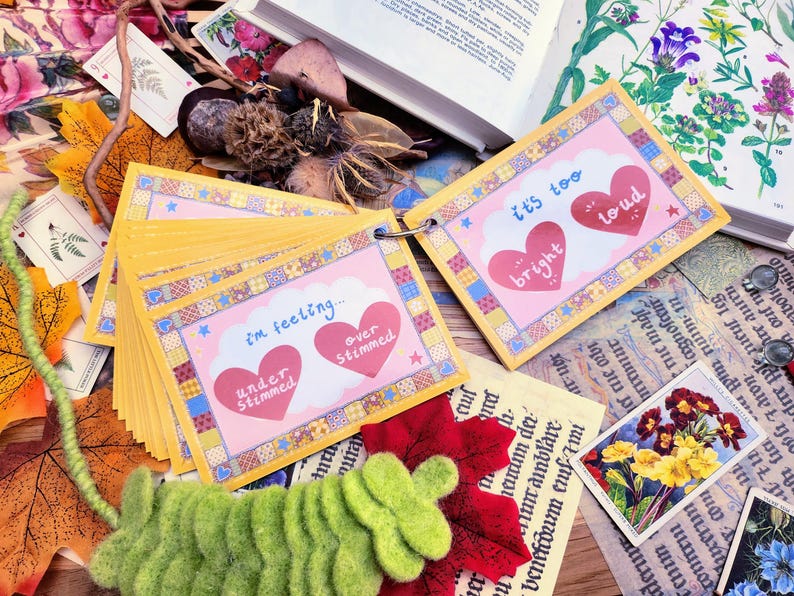 May include: A set of colorful cards with different phrases about feelings, including "I'm feeling... under stimulated, over stimulated", and "It's too bright, it's too loud". The cards are arranged in a stack and have a pink and yellow color scheme.