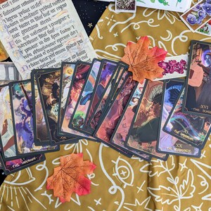 May include: A spread of tarot cards on a gold patterned fabric with fall leaves. The cards are black with gold trim and feature colorful illustrations. The cards include The Unknown, The Dayorean, The Empress, and The Scholar.