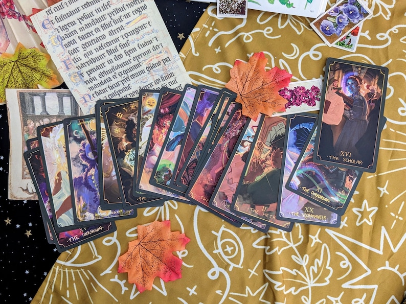 May include: A spread of tarot cards on a gold patterned fabric with fall leaves. The cards are black with gold trim and feature colorful illustrations. The cards include The Unknown, The Dayorean, The Empress, and The Scholar.