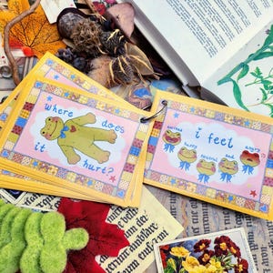 May include: A set of colorful cards with a frog illustration on each card. The cards are bound together with a ring. The text on the cards reads "Where does it hurt?" and "I feel".