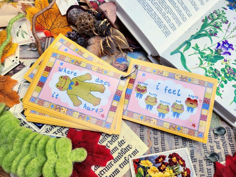 May include: A set of colorful cards with a frog illustration on each card. The cards are bound together with a ring. The text on the cards reads "Where does it hurt?" and "I feel".