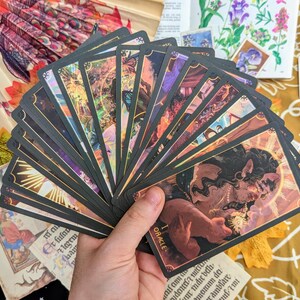 May include: A spread of tarot cards with black and gold borders, featuring illustrations of people and animals. The cards are fanned out in a hand, revealing the top card, which reads "The Oracle".