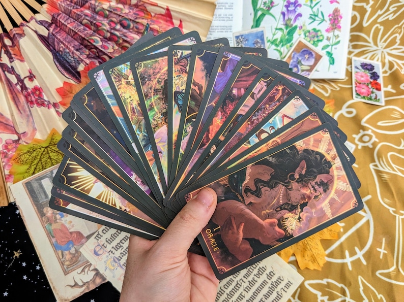 May include: A spread of tarot cards with black and gold borders, featuring illustrations of people and animals. The cards are fanned out in a hand, revealing the top card, which reads "The Oracle".