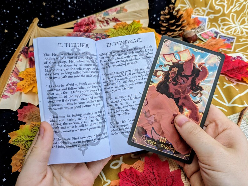 May include: Open book with a colorful illustration of a pirate on the cover. The book is titled "The Pirate" and has text about the meaning of the card. The book is open to a page titled "The Heir" with text about the meaning of the card.