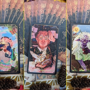 May include: Three tarot cards with black borders and gold accents. The first card shows a person with a rainbow halo holding a glowing orb. The card is titled "The Light". The second card shows a person with red hair and a beard holding a bird. The card is titled "The Inventor". The third card shows a person with green skin and white hair holding a flower. The card is titled "The Witch".