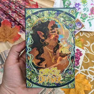 May include: A card from the "The Oracle Diviner" deck features an illustration of a woman with horns and a crown, holding a cup with a glowing orb inside. The card is surrounded by a decorative border of flowers and leaves.
