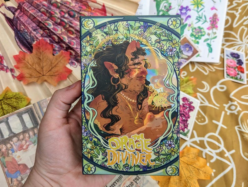 May include: A card from the "The Oracle Diviner" deck features an illustration of a woman with horns and a crown, holding a cup with a glowing orb inside. The card is surrounded by a decorative border of flowers and leaves.
