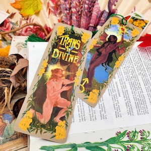 Puede incluir: Dos marcadores ilustrados con detalles dorados. Un marcador presenta una figura desnuda con alas y el texto "TRANS IS DIVINE". El otro marcador presenta una figura desnuda con un halo y el texto "TRANS IS DIVINE".