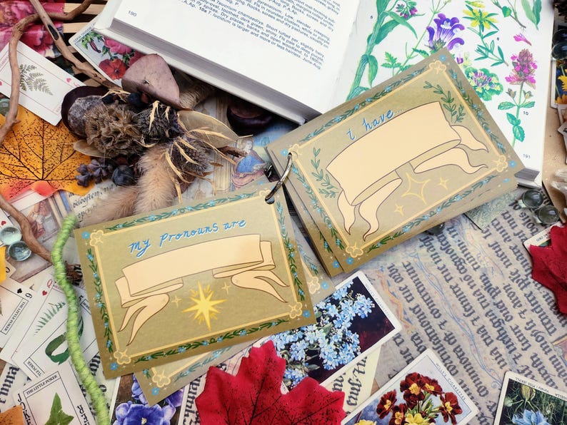 May include: Two small, brown, card-like objects with a gold border and a white banner. The banner on the top card says "I have" and the banner on the bottom card says "My pronouns are".
