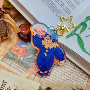 May include: A colourful, glittery, blue and orange character keychain with the text "GOING TO COMMIT ARSON!" in a speech bubble. The character is wearing a blue and white jester costume and has a star-shaped key ring.