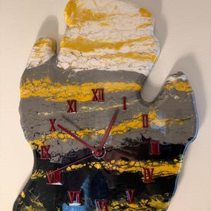 May include: A wall clock shaped like the state of Michigan featuring a fluid acrylic pour design in yellow, white, gray, and black. The clock face displays red Roman numerals and matching red clock hands centered on the abstract painted surface.