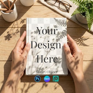 Op de afbeelding: Een hardcover boek met de tekst "Your Design Here" op de omslag, vastgehouden door handen. De omslag heeft een dambordpatroon. De woorden "Smart Object" en drie app-logo's staan onderaan. De achtergrond omvat een houten oppervlak en een kleine potplant.