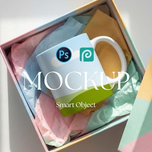 May include: A white ceramic mug with a light blue sky design, green grass, and the word "MOCKUP". The mug features the Adobe Photoshop and Adobe Premiere Pro logos. The mug is inside a colorful gift box with tissue paper.