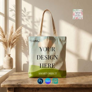 May include: A canvas tote bag with a light blue and green design, featuring the text "YOUR DESIGN HERE" and "SMART OBJECT". The bag has beige handles and is displayed on a wooden table.