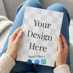 May include: A white book with the text "Your Design Here" in a bold, black font. The book has a checkered pattern on the cover and is held by a person wearing a white sweater and blue jeans. The book cover also includes the words "Smart Object" and the logos for Photoshop, Canva, and Procreate.