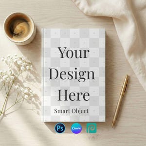 Peut inclure: Maquette de livre avec le texte "Your Design Here" et "Smart Object" sur la couverture. Un stylo doré, une tasse de café et des fleurs blanches sont disposés autour du livre sur une surface en bois clair. La couverture du livre a un motif quadrillé.
